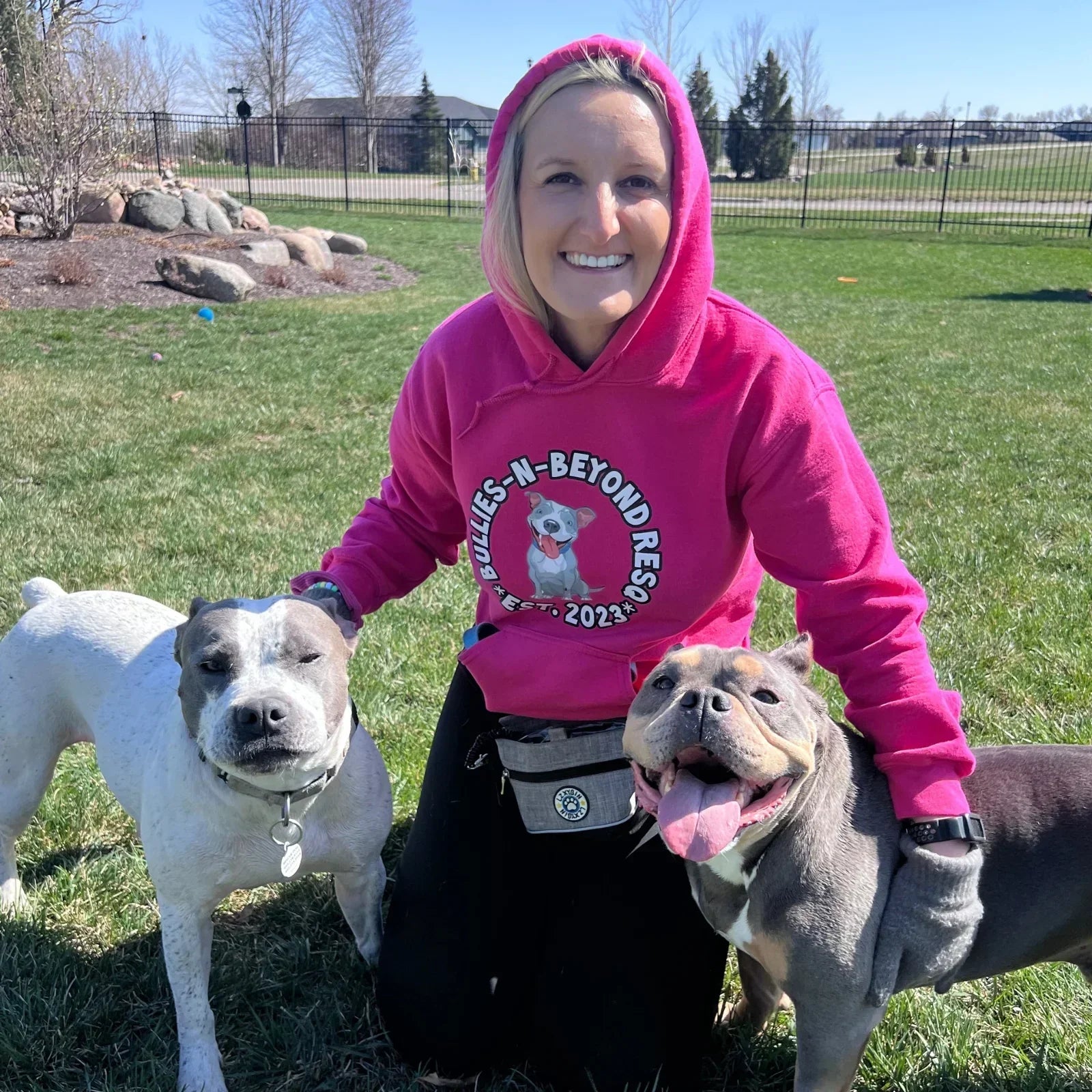 Woman wearing a pink hoodie that says Bullies-N-Beyond ResQ while sitting with two dogs in the yard.