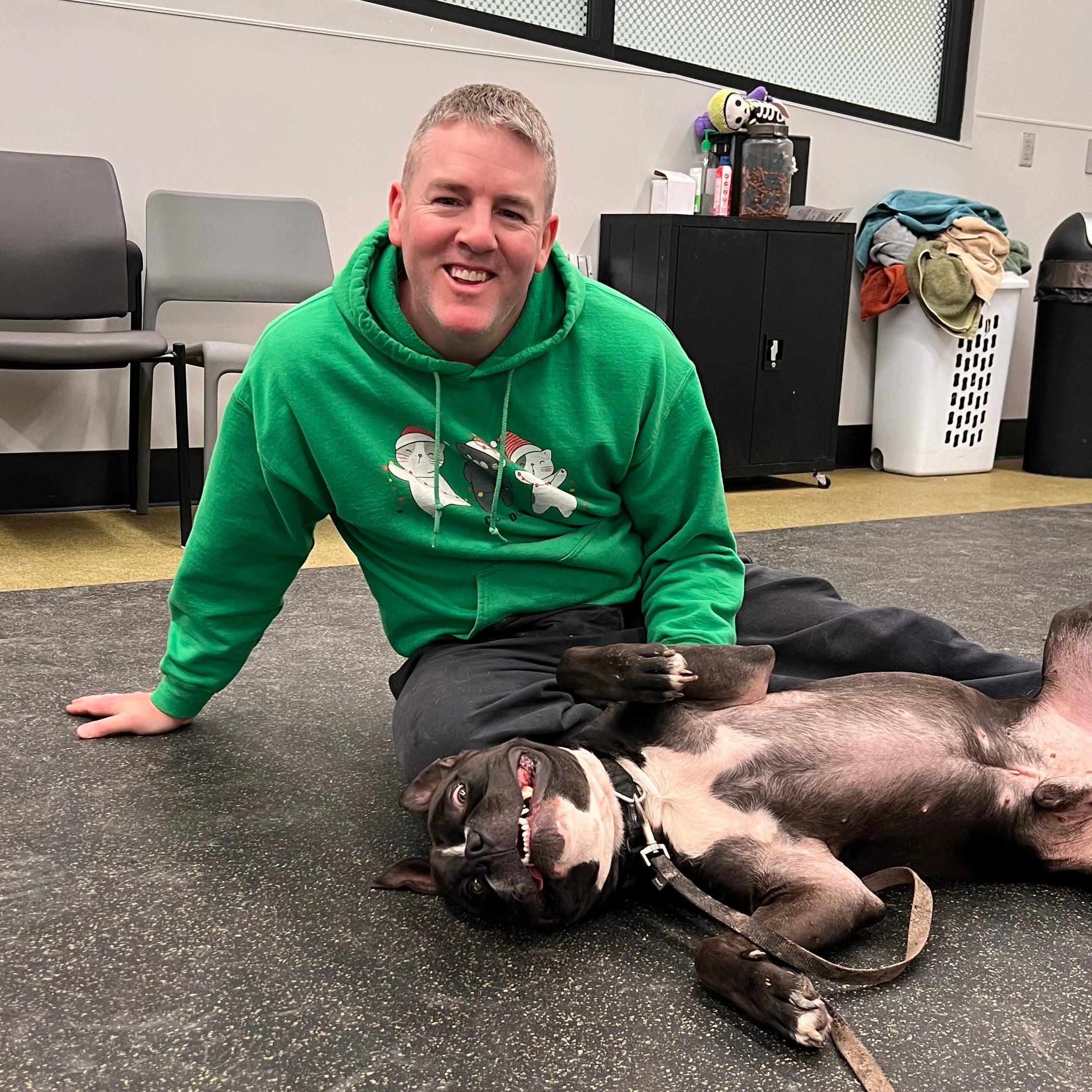 Man in a green hoodie with a dog on a carpeted floor in an indoor setting